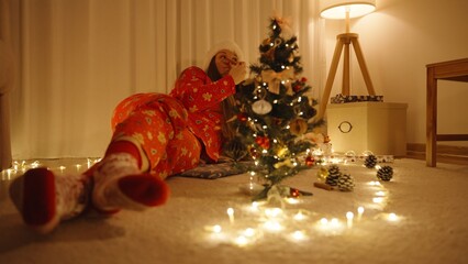 Woman in Festive Pajamas Lying on Floor and Decorating Mini Tree Surrounded by Warm Lights and Pinecones, Low Angle, Cozy Christmas Concept
