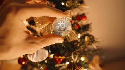 Hand Holding Bitcoin in Front of Decorated Tree with Lights and Ornaments, Close Up View, Cryptocurrency Christmas Concept