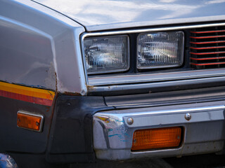 The front corner of a silver classic car from the 1980s showing the double headlights and amber turn signal The chrome bumper and boxy design are iconic elements of this retro automobile
