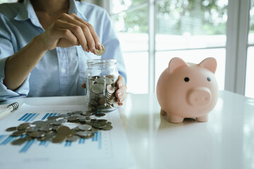 A person places a coin into a pink piggy bank, symbolizing saving money, financial planning,...