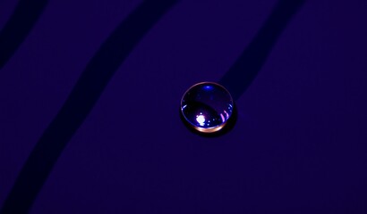 A single, reflective water droplet rests on a deep blue, subtly textured surface. Dark lines intersect behind it, creating a dramatic contrast.