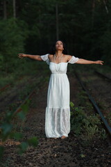 Beautiful dark haired curly woman in long white dress standing on the railway in the forest in the...