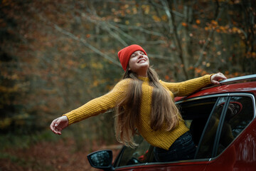 Portrait of a teenage girl in the window of a red car against the background of an autumn forest. Enjoying life.