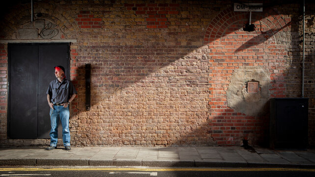 A Working Relationship. A man in a casual outfit with a red cap stands in front of a brick wall, bathed in a strong, diagonal beam of sunlight.