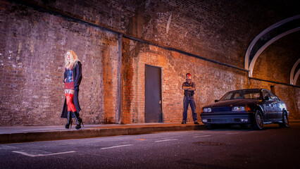 A Working Relationship. A sex worker stands waiting for business on a city pavement at night. Her guardian is in the background acting as protection. From an image series featuring character models.