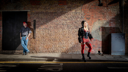 A Working Relationship. A sex worker stands waiting for business on a city pavement at night. Her guardian is in the background acting as protection. From an image series featuring character models.