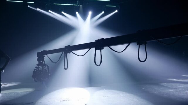 Professional camera and stabilizer rig hanging from a camera crane, backlit by dramatic converging white spotlights and fog on an empty stage, conveying cinematic production