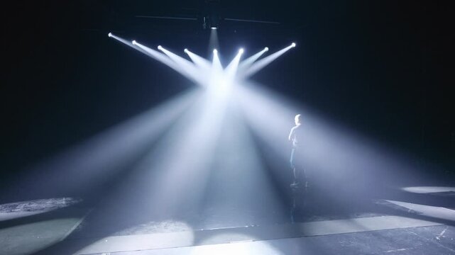 Atmospheric view of a stage dramatically illuminated by multiple converging white spotlights, with beams cutting through fog and the silhouette of a person standing, conveying a powerful show