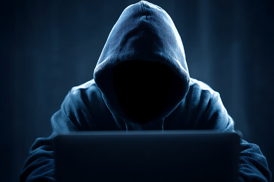 Hooded hacker committing cyber crime using laptop in dark room