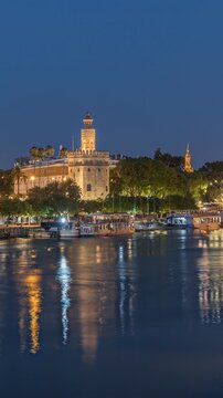 Torre del Oro watchtower in Seville, Spain. Day to night transition timelapse with city lights reflecting on water and scenic aerial view of waterfront. Tour boat floating along the Guadalquivir River