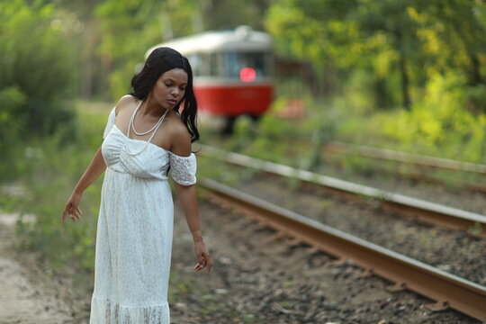 Beautiful young dark haired curly woman in white dress standing near rails and red tram in the forest