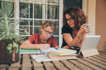 Young student doing homework at home with school books newspaper, digital pad helped by his mother. Mum control, help and teaching his son. Education, family, lifestyle and homeschooling concept.