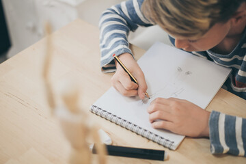 Student drawing with pencil a human figure be inspired by a wooden model. Boy copying a dummy for school art tasks. Kid hold a pencil and draw a manga at home. Education, talent and ability concept. © Fabio Principe