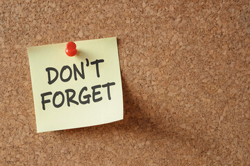 Yellow sticky note with D O N' T F O R G E T text pinned to corkboard paper dont forget