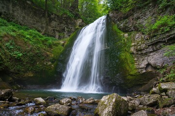 A peaceful cascade of water tumbling over rugged rocks, enveloped by vibrant greenery and moss in a woodland setting.