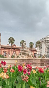 Hispalis Fountain in Seville, one of the city's most beautiful fountains, surrounded by colorful flowers and historical buildings. Timelapse hyperlapse with a cloudy sky in the background.