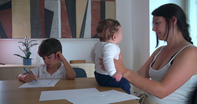 Boy working on homeschooling assignments at the table while mother cradles baby, demonstrating concentration and family-oriented homeschooling in a peaceful home setting focused on education