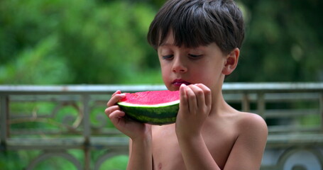 Boy deeply focused on eating a slice of watermelon, enjoying the juicy fruit outdoors on a balcony, savoring the moment in a peaceful and relaxed setting