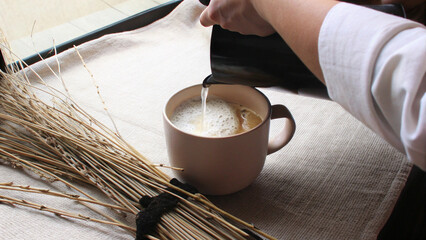 Barista in white shirt pouring hot water into beige coffee mug with foam on linen tray, clean top view cafe setup for warm drink preparation in soft natural morning light