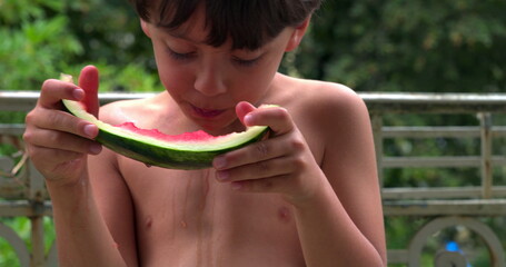 Young boy carefully eating a slice of watermelon, dripping juice as he enjoys the fresh and sweet...