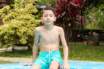 Portrait of a Hispanic boy with his eyes closed sitting on the edge of a hotel pool in Rivera,...