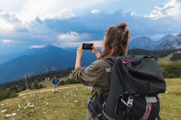Young woman with a backpack photographing a beautiful misty mountain panorama with her smartphone during a recreational hiking tour.