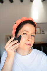 Middle aged woman with a pink headband gently glides a black gua sha tool across her cheek....