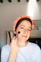 Confident middle aged woman doing her makeup with a brush and looking at camera, in a bright and...