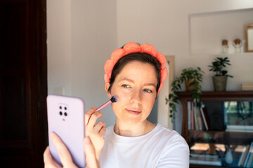 Middle-aged woman holding a smartphone and looking at screen while filming a beauty tutorial in a...