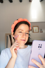 Middle aged woman in a white shirt and pink headband gently rolls a facial tool on her cheek. She...