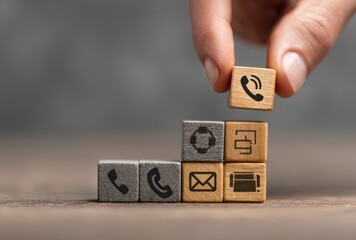 Hand places block showing phone symbol atop stack of wood blocks, featuring various contact & service icons, on a textured background