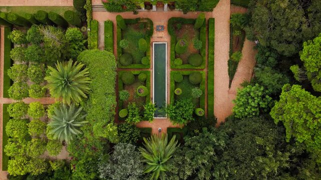 Drone video captures a public garden in Spain, showing symmetrical landscaping, lush greenery, palm trees, and a pool beside a traditional building.
