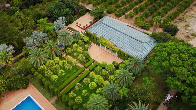 Drone video shows a lush public garden in Spain, with structured greenery, a pool, and architectural elements surrounded by orchards and scenic landscapes.