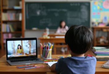 Young student virtually attends class via laptop. School desk is filled with pencils, crayons, books; chalkboard/teacher behind student