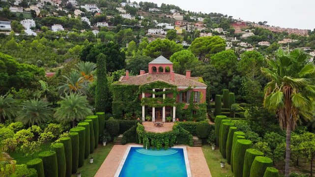 Drone video of a Spanish public garden featuring a historic ivy-covered building, elegant topiary, palm trees, and a pool surrounded by lush greenery.