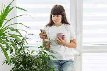 Woman reading phone while holding coffee indoors. Morning routine