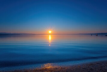 Sunburst over a calm, still lake, reflecting light and colors. Distant shoreline is visible. Waves lap gently at the pebbled shore in the foreground