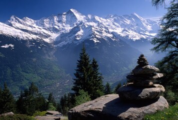 Scenic alpine vista shows snowy mountains, green valley, and cairn of balanced rocks on a boulder in the foreground, lit with sunlight