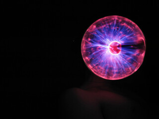 A guy holds a red and blue plasma ball with rays in the dark next to his head	
