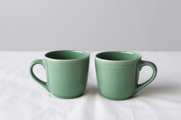 A pair of green ceramic mugs rest side by side on a white fabric, showcasing a minimalist and graceful style.