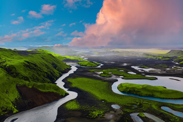 Aerial view of Iceland's landscape with green moss covered hills, winding rivers, scattered lakes, volcanic soil, and a faint rainbow in the cloudy sky.