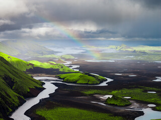 Aerial view of Iceland's green moss covered hills, dark volcanic plains, winding river, scattered lakes, and a faint rainbow on the horizon.