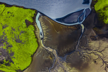 Aerial image of Iceland's green moss covered terrain, dark volcanic soil, and a pale winding river flowing toward a serene lake with tributary patterns.