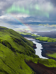 Aerial view of green moss covered hills, a white river cutting through dark volcanic terrain, and a faint rainbow under a cloudy Icelandic sky.