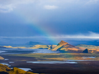 Aerial view of rugged Icelandic landscapes featuring volcanic craters, scattered lakes, a mountain peak, a faint rainbow, and dramatic cloud cover.