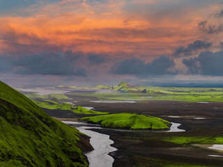 Aerial view of Iceland's green moss covered hills, dark volcanic soil, and a winding river. A faint rainbow arcs over the untouched natural landscape.