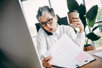 Mature businesswoman multitasking with documents, phone call, coffee, and computer in a contemporary office environment