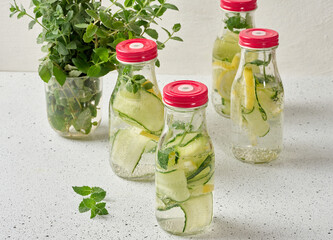 Glass bottles with red lids, filled with detox water containing cucumber and lemon slices and mint leaves, white table