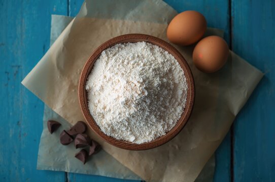 Wooden bowl filled with flour, eggs, chocolate chips, and baking parchment on a blue surface. Overhead shot