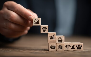 Hand stacks wooden cubes with business icons on a wood surface against a blurred dark background